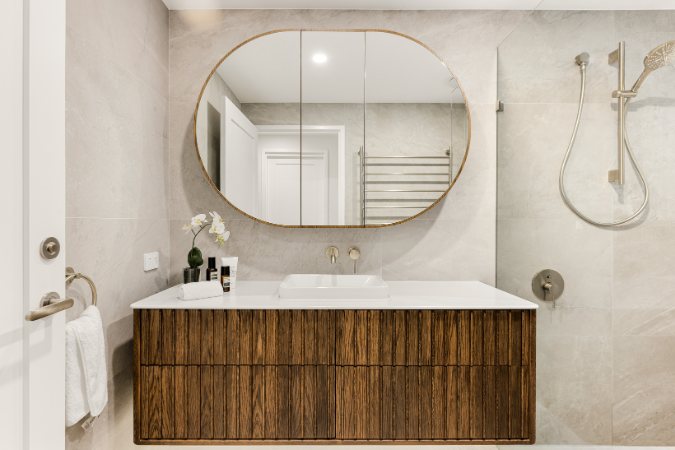 Bathroom vanity with an oval mirror, fluted timber cabinetry and a handheld shower beyond — Kangaroo Point Villa 8