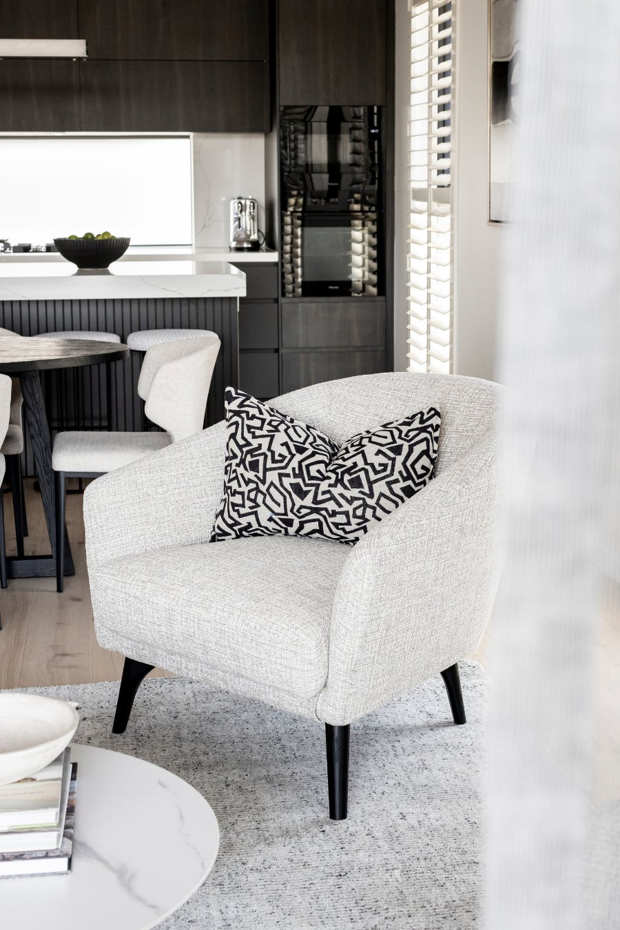 Living area armchair with a patterned cushion, dark kitchen cabinetry and dining beyond — Monochrome Residence