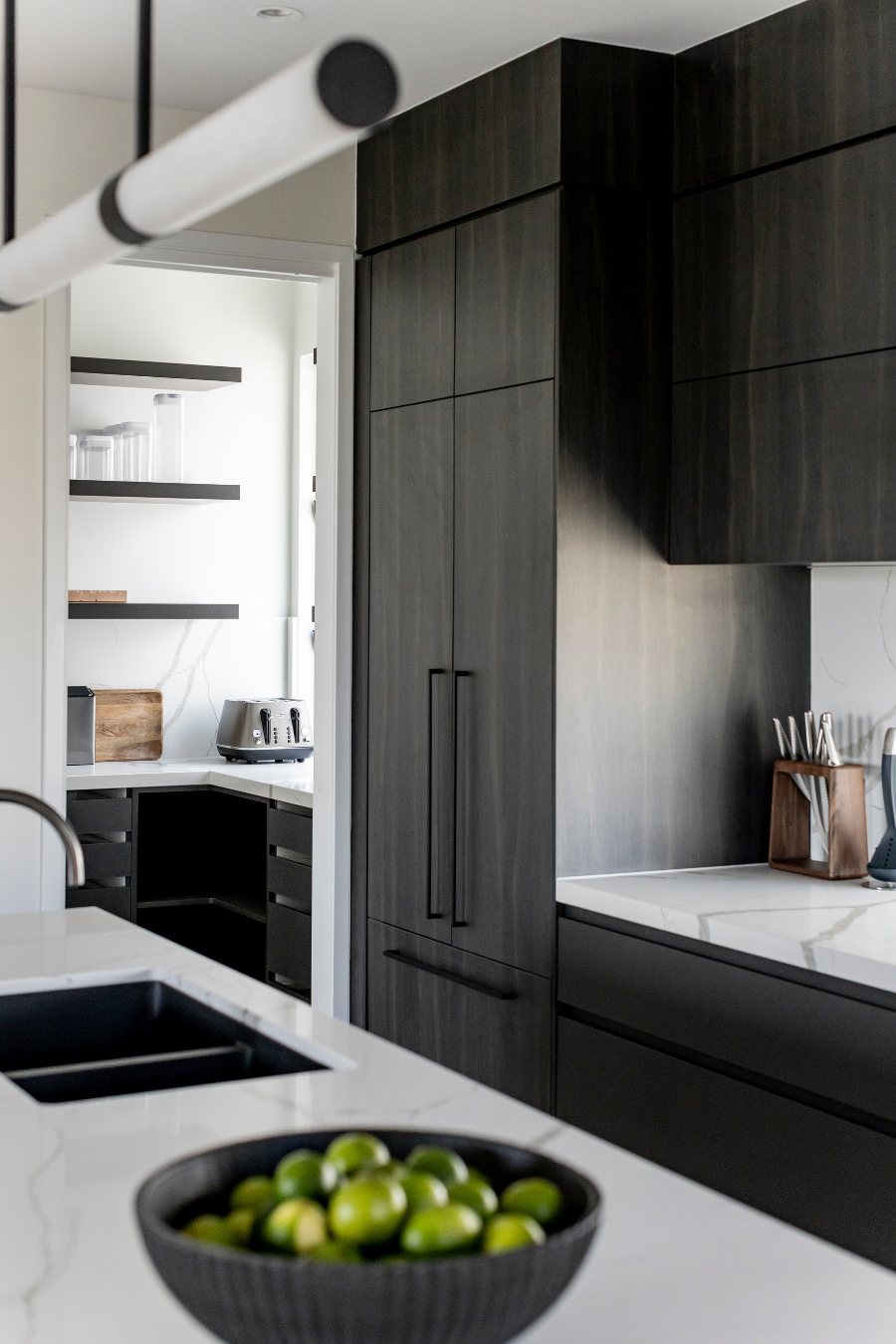 Dark timber kitchen cabinetry with a butler’s pantry visible beyond and a bowl of limes on the island — Monochrome Residence