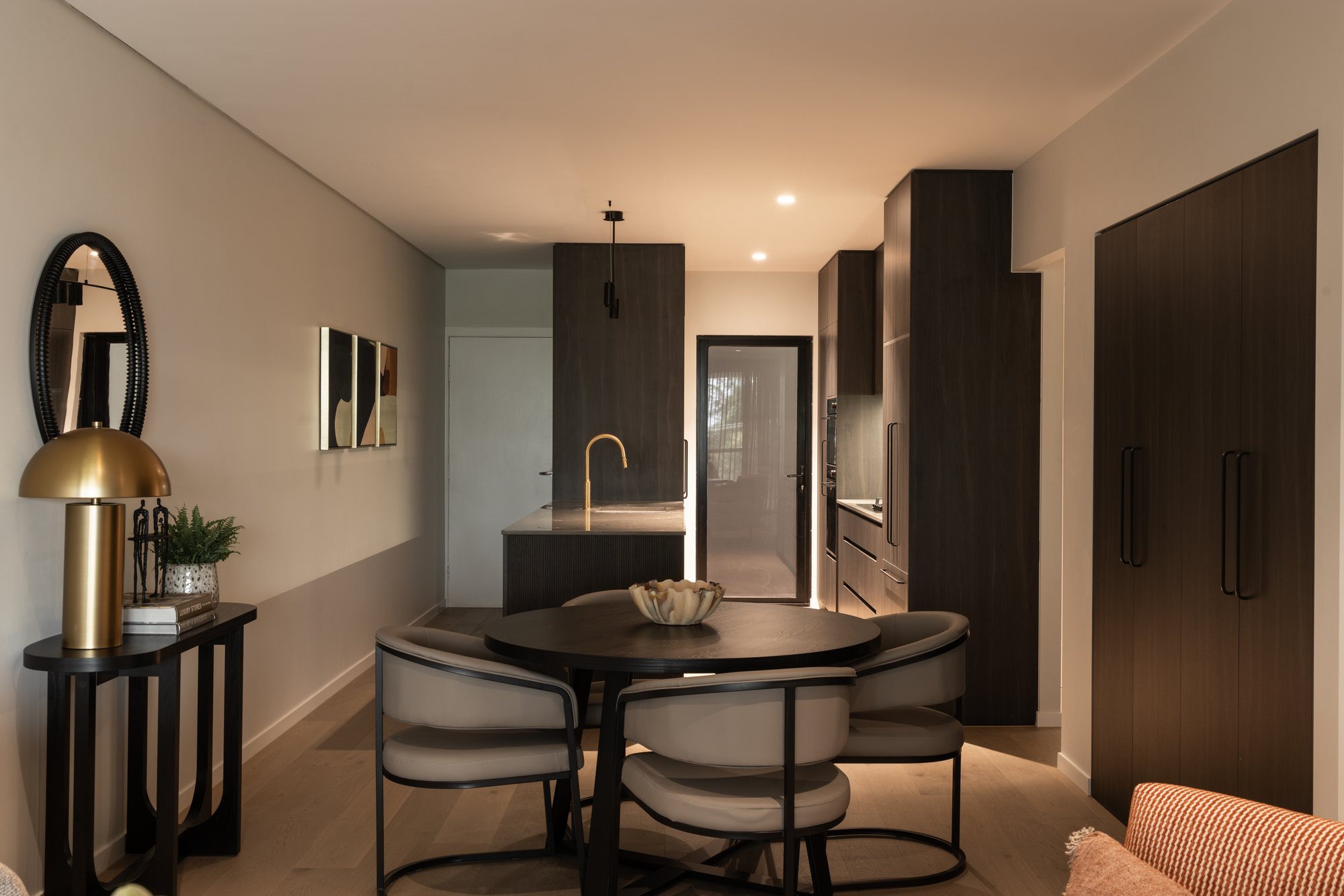 Dining nook with a round table and sculptural chairs beneath a brass pendant, dark kitchen cabinetry beyond — Nundah