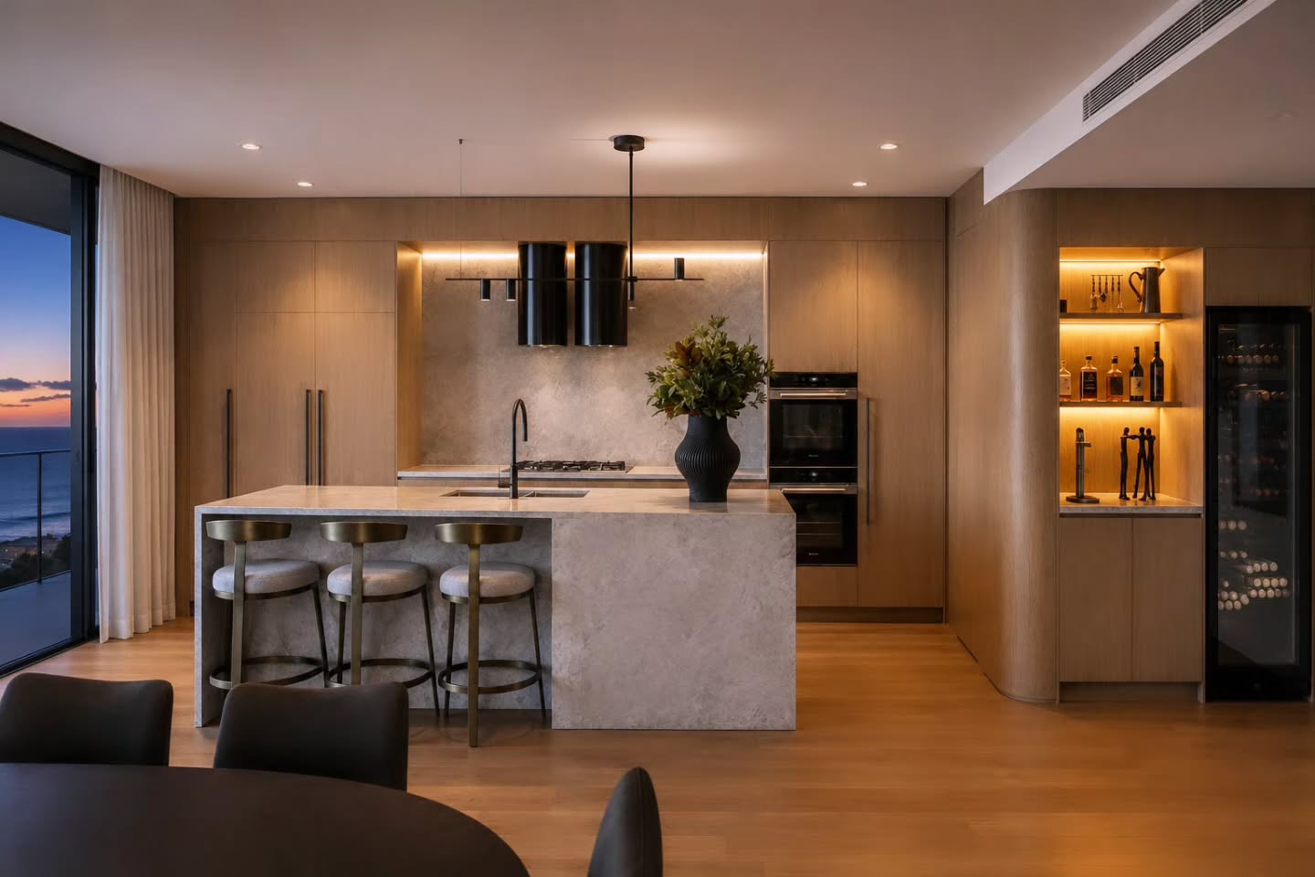 Timber kitchen at dusk with a stone island, black pendant lights and wine display, ocean beyond — Oceanfront Apartment