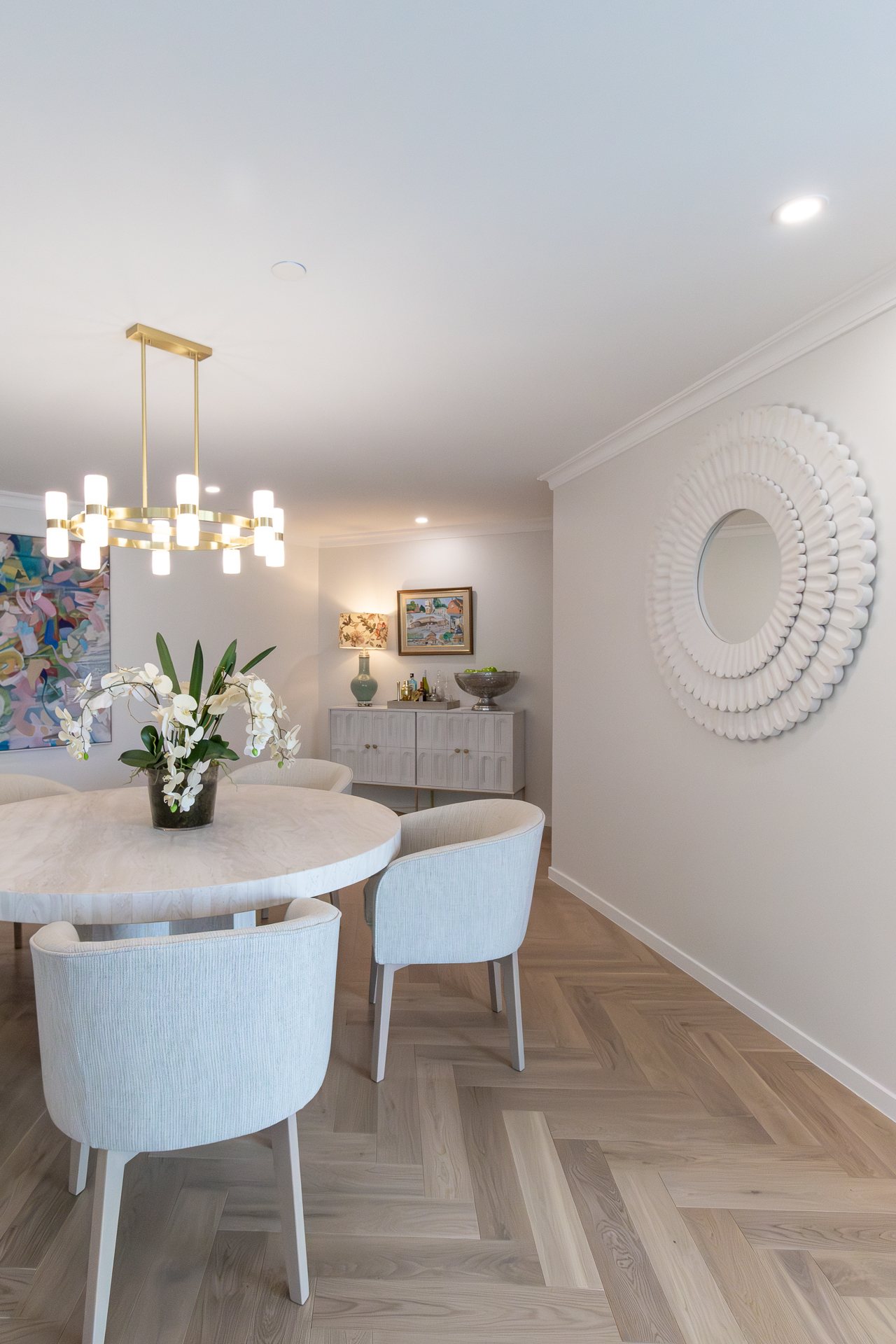 Round marble dining table beneath a brass chandelier, with a sunburst mirror and credenza beyond — Rivage