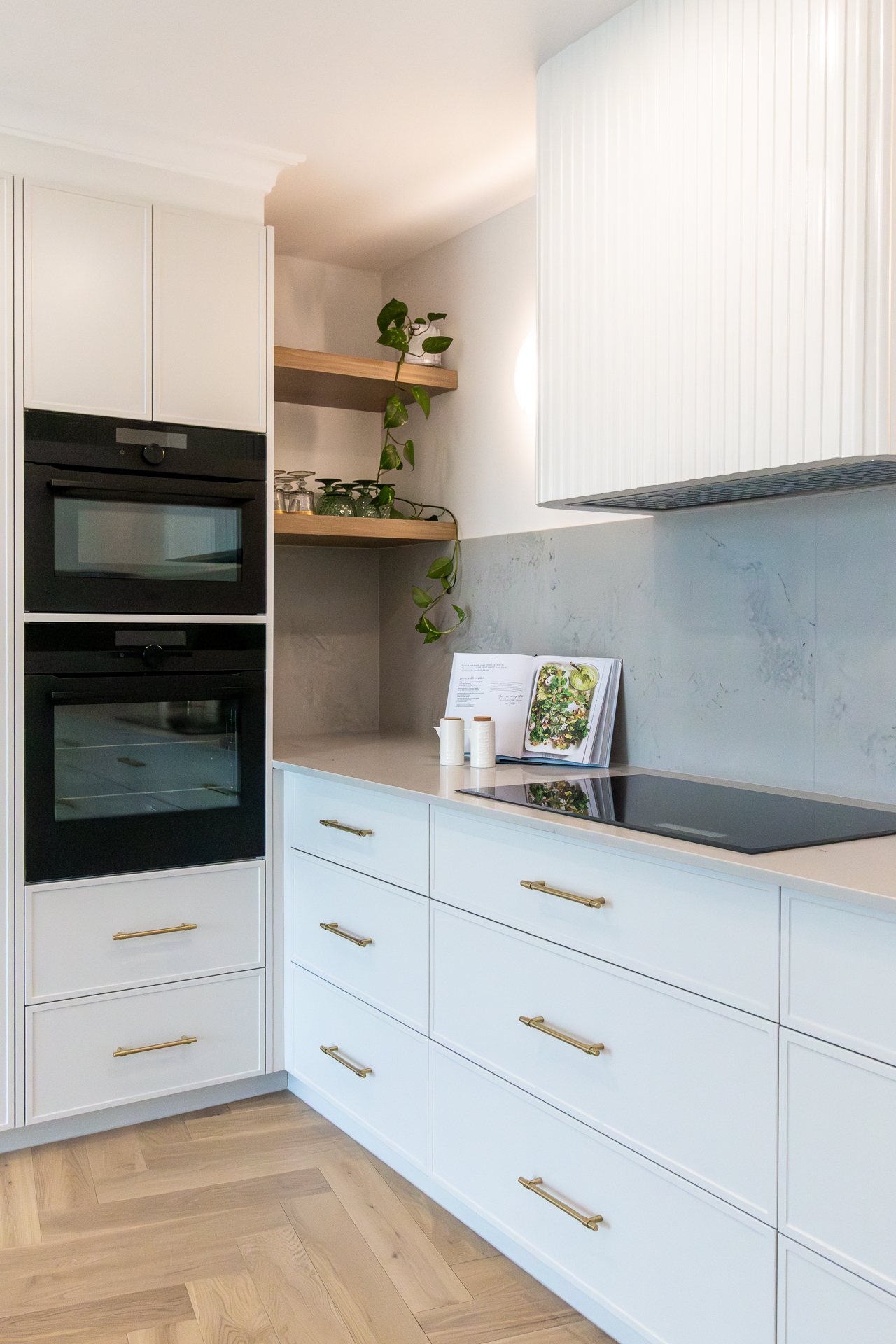 Kitchen with white cabinetry, black built-in ovens and open timber shelving — Rivage