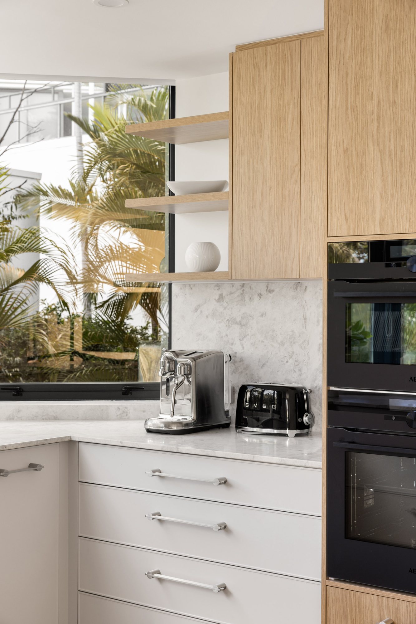 Floating oak shelving and marble splashback beside a window onto palms — Stradbroke Villa 8