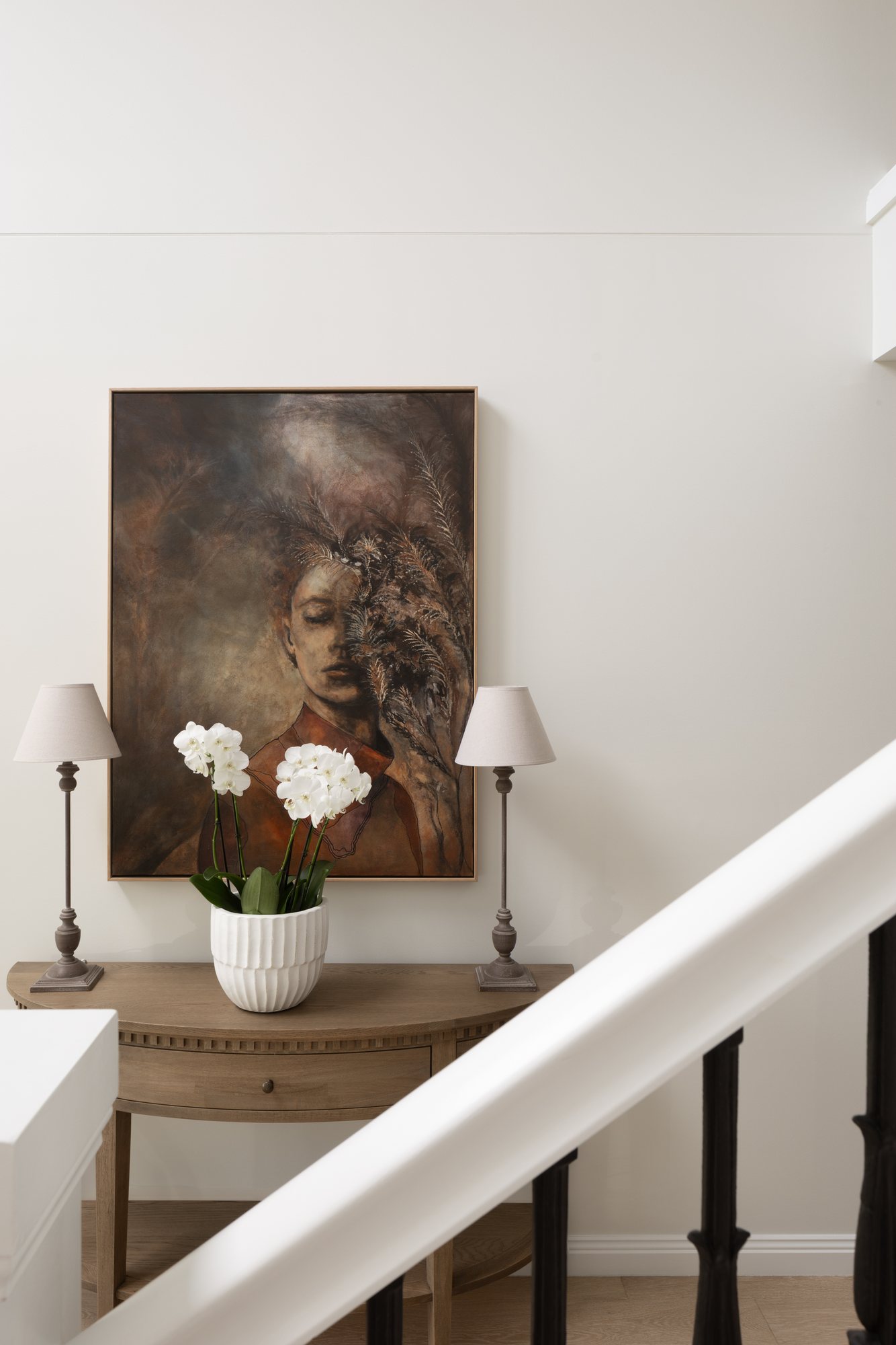 Stairwell console table with a framed portrait artwork, table lamps and orchids — The Manor House